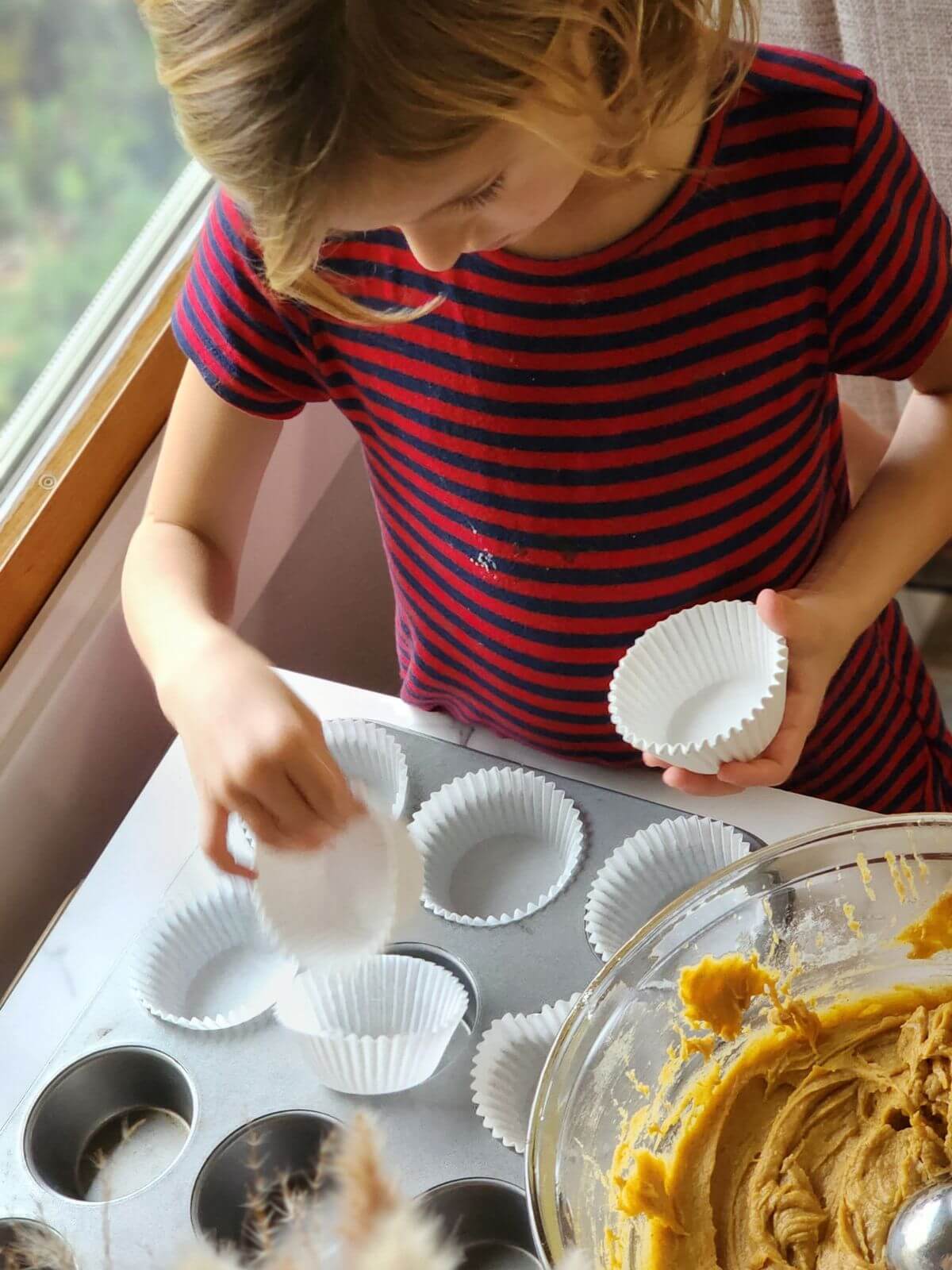 Child placing paper liners into a muffin pan beside a bowl of pumpkin cupcake batter.