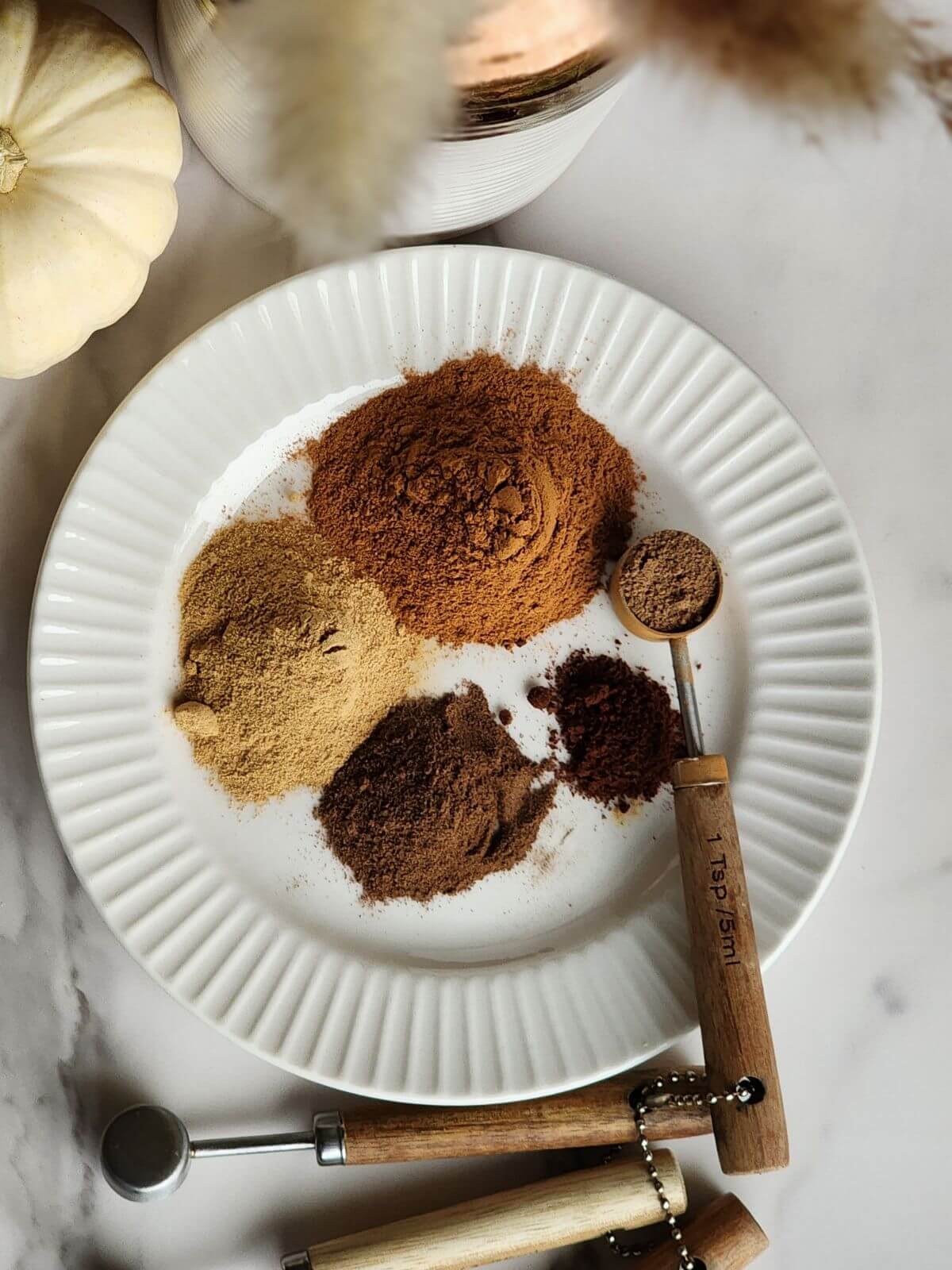 Pumpkin pie spices measured onto a white plate beside wooden measuring spoons.