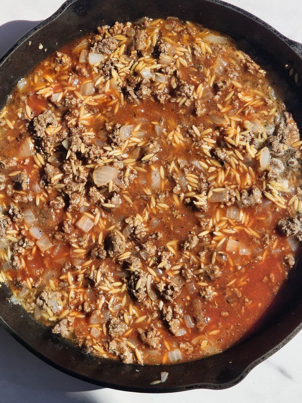 Tomato sauce and orzo added to browned ground beef and onions in a cast iron skillet.