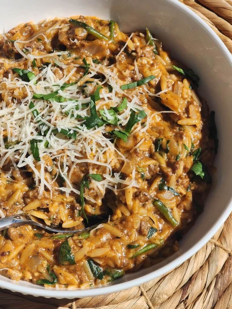 Creamy ground beef orzo with tomato sauce, parmesan cheese, spinach, and fresh basil in a white serving bowl.