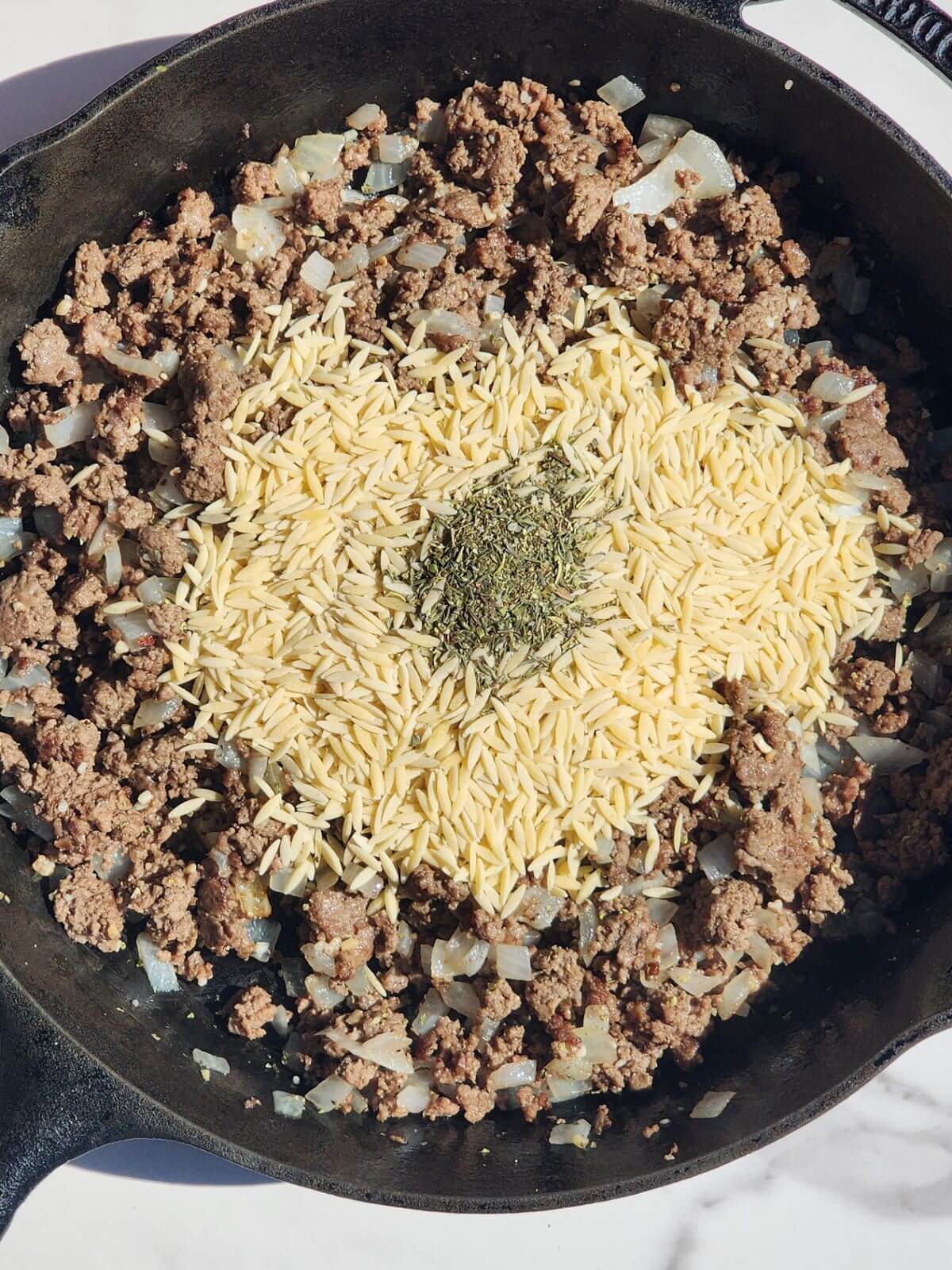 Dry orzo and Italian seasoning added to browned ground beef and onions in a cast iron skillet.