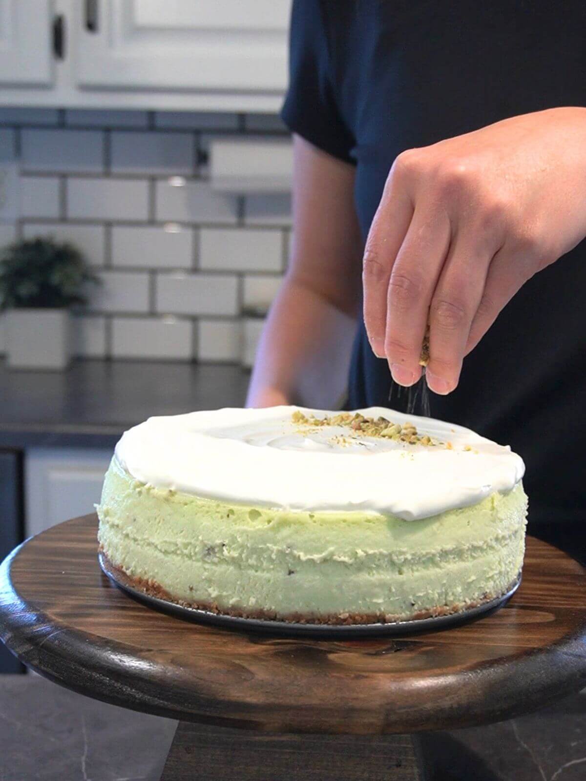 Chopped pistachios being sprinkled over pistachio cheesecake with sour cream icing.