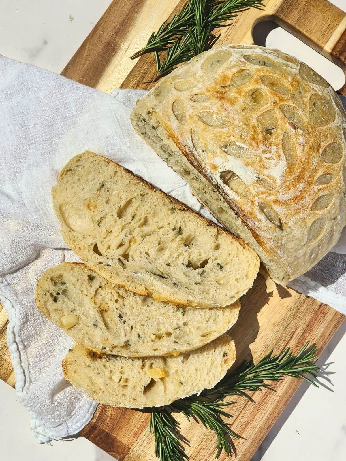 Sliced roasted garlic and rosemary sourdough bread served on a wooden board with fresh rosemary.