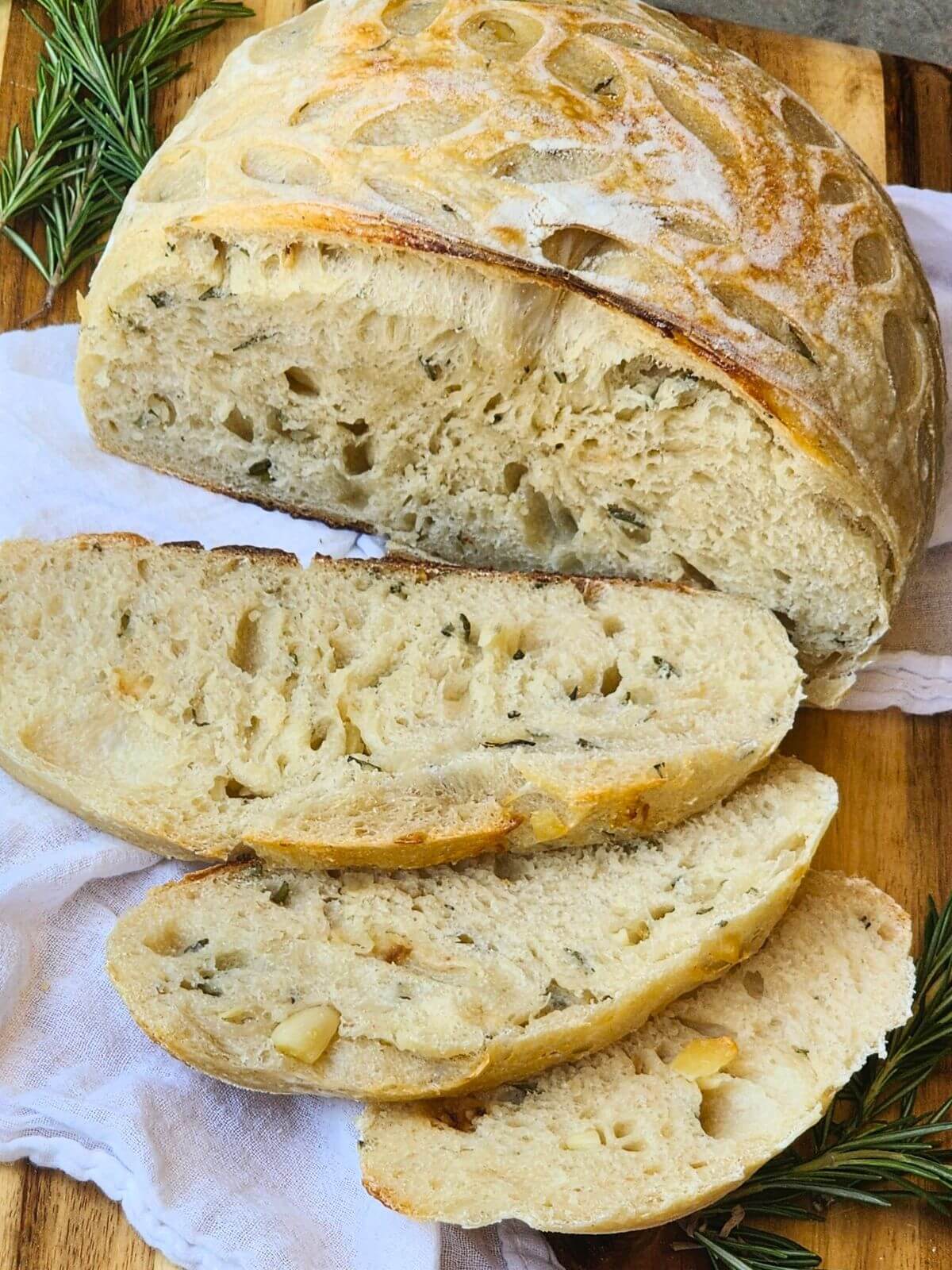 Sliced roasted garlic and rosemary sourdough bread served on a wooden board with fresh rosemary.