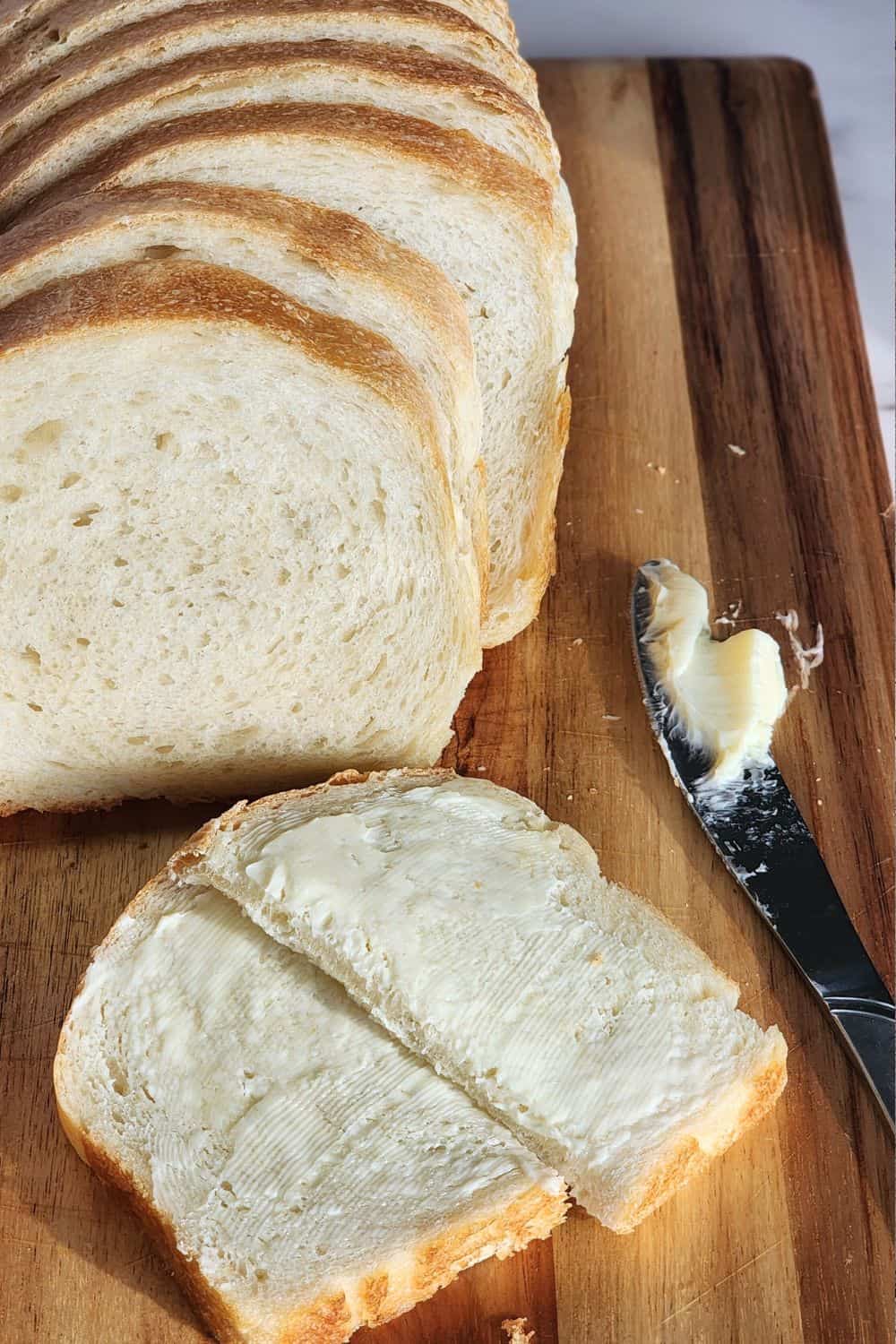 Loaf of sourdough sandwich bread with a buttered piece laying on cutting board along with a knife with butter on it.
