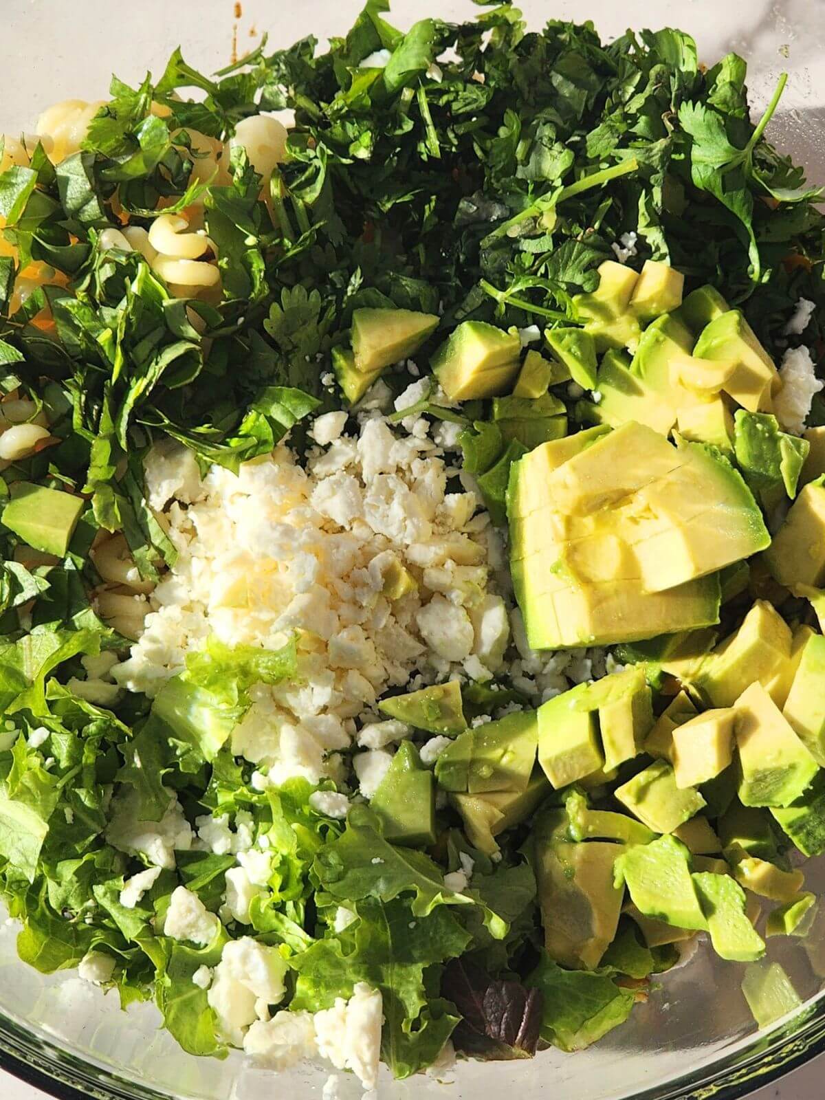 Avocado, cotija cheese, cilantro, and basil added to a bowl of pasta, corn, and lettuce for Mexican street corn pasta salad.