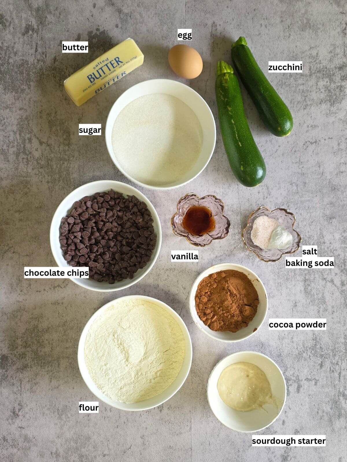Ingredients for sourdough chocolate chip zucchini brownies, including butter, sugar, egg, zucchini, chocolate chips, flour, cocoa powder, vanilla, sourdough starter, salt, and baking soda.