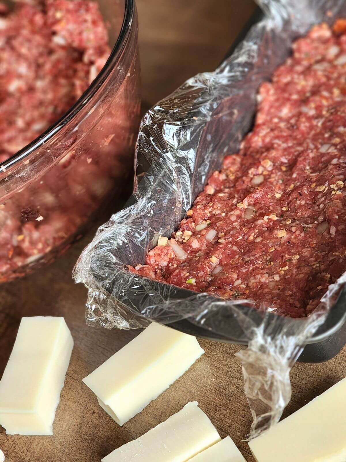 Ground beef mixture pressed into a plastic lined loaf pan with slices of cheese ready for smoked meatloaf.
