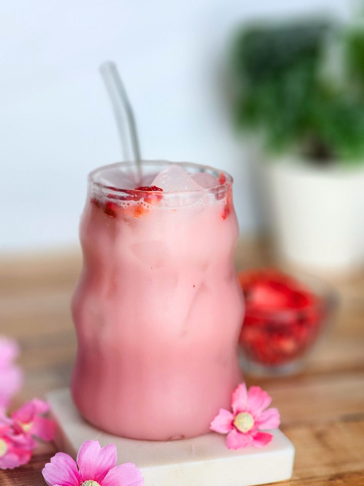 A creamy pink drink served over ice in a clear glass with freeze dried strawberries on top and pink flowers around the glass.