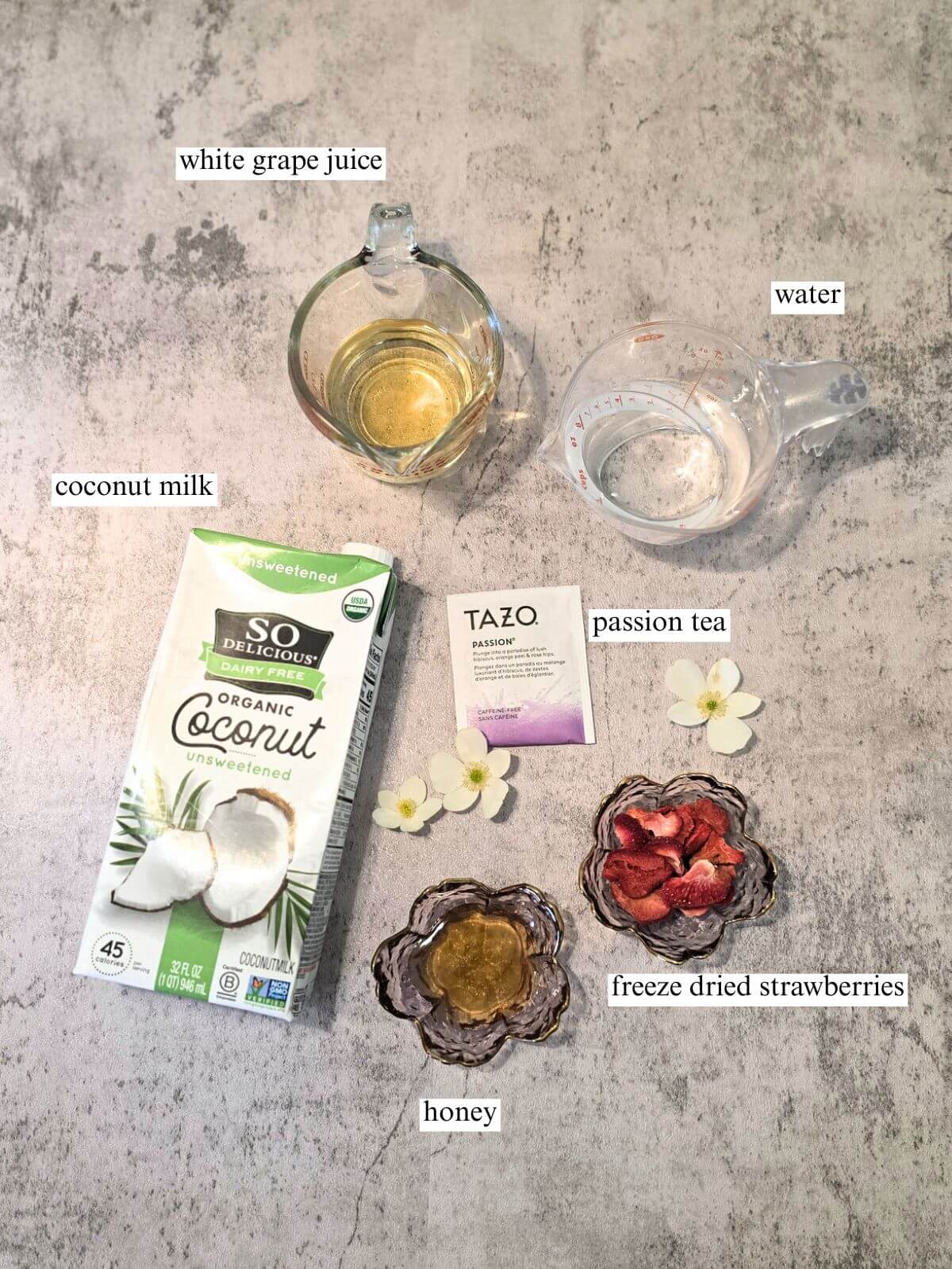 Labeled ingredient photo for a homemade pink drink with Passion tea, white grape juice, coconut milk, honey, freeze dried strawberries, and water arranged on a gray backdrop.