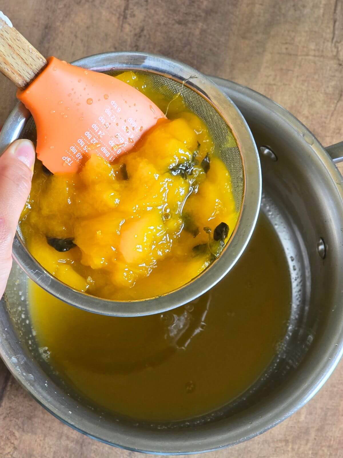 Straining cooked mango and basil through a fine mesh sieve to make mango syrup.