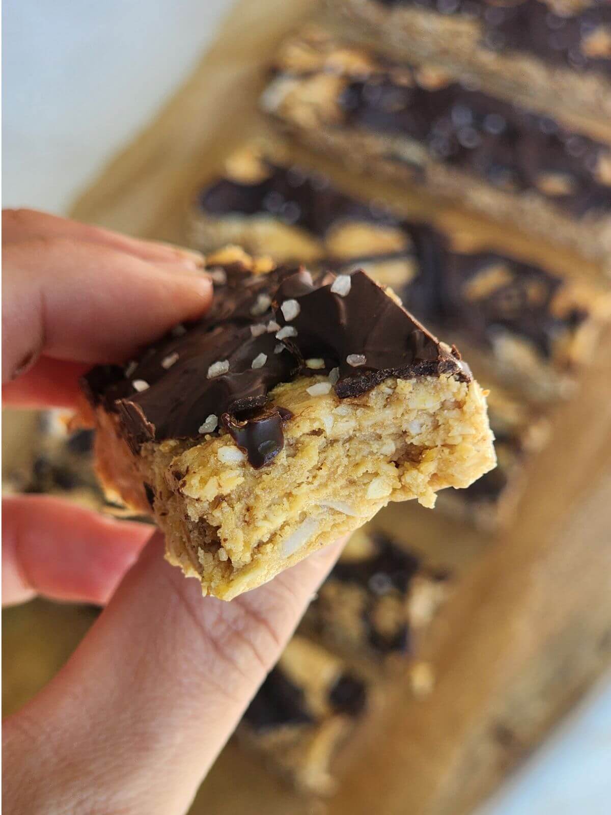 Hand holding a chocolate coconut protein bar topped with melted chocolate and flaky sea salt.