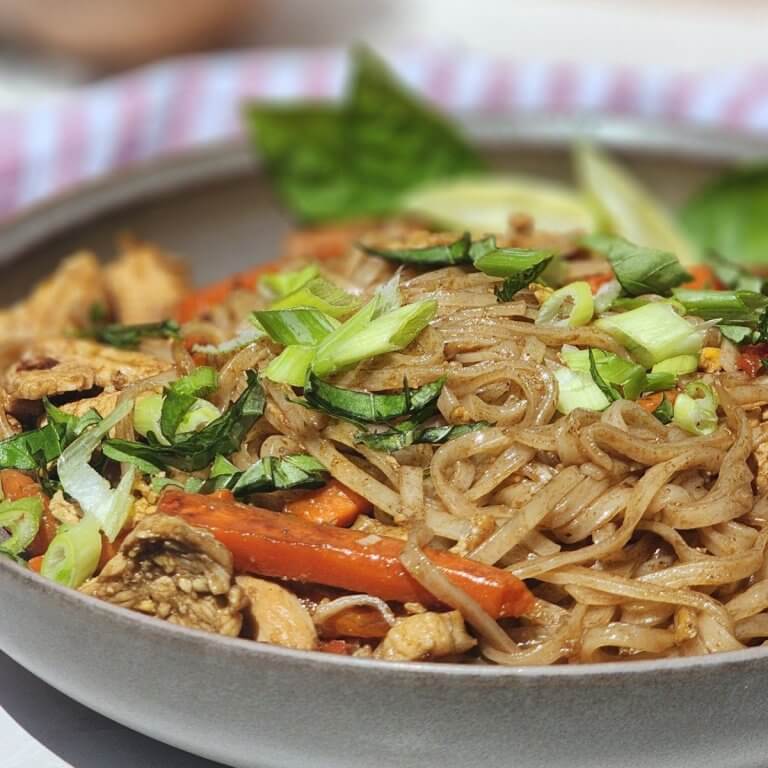 Easy chicken Pad Thai served in a bowl with chicken, rice noodles, carrots, red bell pepper, green onions, basil, and lime.