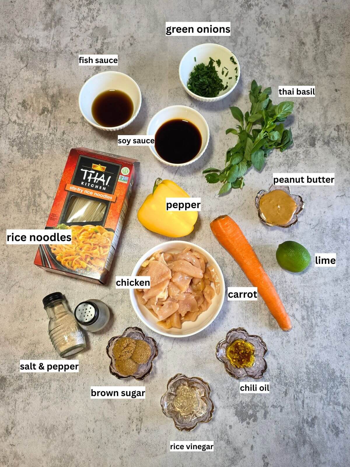 Ingredients for easy chicken Pad Thai, including chicken, flat rice noodles, bell pepper, carrots, green onions, garlic, lime, soy sauce, fish sauce, rice vinegar, peanut butter, honey, and chili oil.